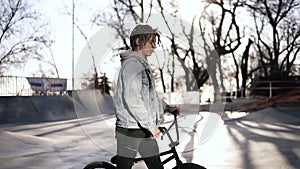 A young boy with short dreadlocks in the skate park wak lking by his bmx bike. Sunny weather. Side view footage