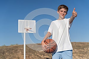 Young boy showing thumb up while posing with basketball ball at an outdoor court.