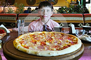 Young boy sitting in front of a large pizza in a cafe. He licked and shows that he is very hungry