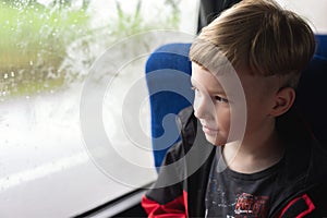 young boy sitting near foggy and misted window in bus or train and looking on raindrops on wet glass