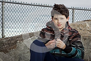 Young Boy Sitting on a Rock in Front of a Chain Link Fence Under a Cloudy Grey Sky