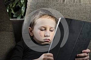 Young boy sitting on sofa in living room and watching cartoons on tablet. Portrait of a pre-school child using devices at home