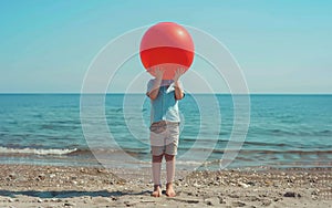 Young boy standing alone holding a red balloon in front of face on the beach