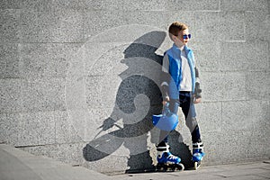 Young boy standing near grey wall with inline roller skates and all protection at outdoor skate park