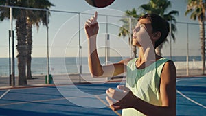Young boy stands and juggles ball on finger on basketball court against blue sky. Basketball player shows skills of