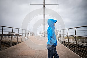 Young boy stood on bridge looking out to sea away from camera hood up in blue coat rain storm weather and wind blowing outdoors