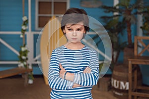 Young boy in the striped shirt stands frowning against the backdrop of the summer house. Studio shot