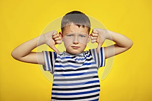 A young boy in striped t-shirt showing thumbs down gesture  with two hands on yellow background. Displeased child giving thumbs