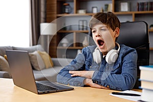 Young boy studying remotely at home using laptop. Tired schoolboy yawning during an online lesson with teacher. Distance