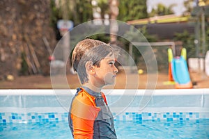 Young boy in swimwear standing contemplatively at poolside with garden in background