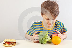 Young boy at the table chooses between hamburger and healthy diet on white background