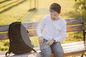 Young boy take out of his pocket smartphone. Teenage boy sitting on the bench in the park