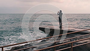 Young boy taking pictures of stormy sea. Man making photo standing at pier. Lonely boy taking video of windy ocean. Side view of y