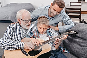 Young boy trying to learn playing guitar while his father and grandpa are