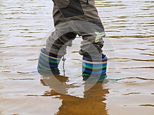 Young boy is washing rubber boots in muddy water of pond. Dirty sand