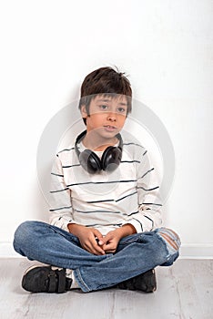 A young boy wearing a white shirt and jeans with headphones siting on the floor by the wall and thinks or sad about something