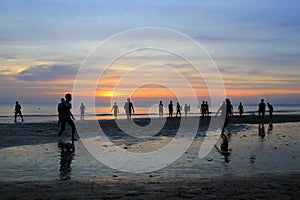 The young boys are playing football on the beach on the background of colorful sunset.