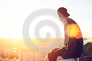 Young and brave man sitting on the edge of the roof and looking far away at the city