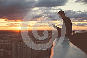 Young brave man sitting on the edge of the roof with smartphone