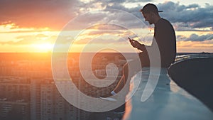 Young brave man sitting on the edge of the roof with smartphone at sunset