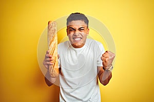 Young brazilian baker man holding bread standing over isolated yellow background screaming proud and celebrating victory and