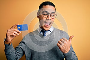 Young brazilian businessman holding credit card money over isolated yellow background pointing and showing with thumb up to the