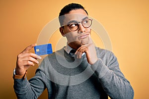 Young brazilian businessman holding credit card money over isolated yellow background serious face thinking about question, very