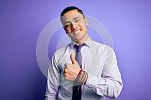 Young brazilian businessman wearing elegant tie standing over isolated purple background doing happy thumbs up gesture with hand