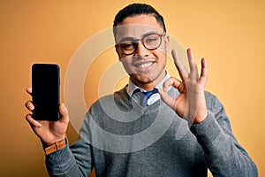 Young brazilian businessman wearing glasses holding smartphone showing screen doing ok sign with fingers, excellent symbol