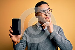 Young brazilian businessman wearing glasses holding smartphone showing screen serious face thinking about question, very confused