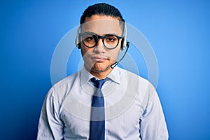 Young brazilian call center agent man wearing glasses and tie working using headset with serious expression on face