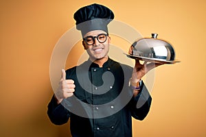 Young brazilian chef man wearing cooker uniform and hat holding tray with dome happy with big smile doing ok sign, thumb up with