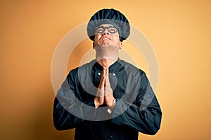 Young brazilian chef man wearing cooker uniform and hat over isolated yellow background begging and praying with hands together