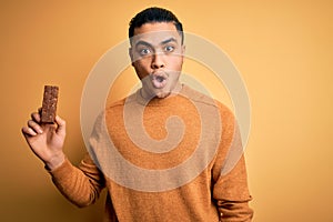 Young brazilian man eating healthy energy bar with protein over isolated yellow background scared in shock with a surprise face,