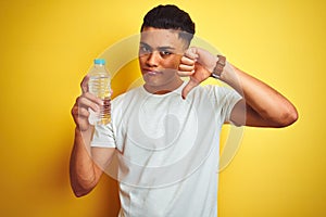 Young brazilian man holding bottle of water standing over isolated yellow background with angry face, negative sign showing