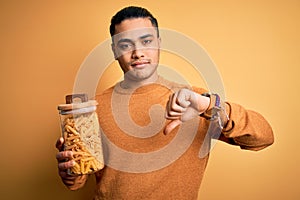 Young brazilian man holding jar with Italian dry pasta macaroni over isolated yellow background with angry face, negative sign