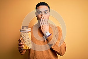Young brazilian man holding jar with Italian dry pasta macaroni over isolated yellow background cover mouth with hand shocked with