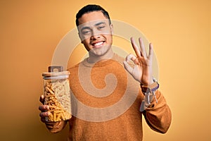 Young brazilian man holding jar with Italian dry pasta macaroni over isolated yellow background doing ok sign with fingers,