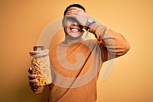 Young brazilian man holding jar with Italian dry pasta macaroni over isolated yellow background stressed with hand on head,