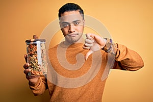 Young brazilian man holding jar with Italian dry pasta over isolated yellow background with angry face, negative sign showing