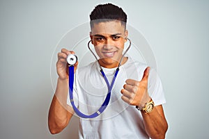 Young brazilian man holding stethoscope standing over isolated white background happy with big smile doing ok sign, thumb up with