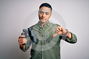 Young brazilian man looking through binoculars over isolated white background with angry face, negative sign showing dislike with