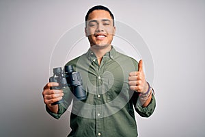 Young brazilian man looking through binoculars over isolated white background happy with big smile doing ok sign, thumb up with