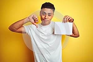 Young brazilian man showing paper banner standing over isolated yellow background with angry face, negative sign showing dislike