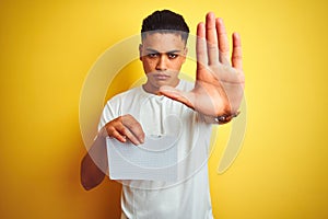 Young brazilian man showing paper banner standing over isolated yellow background with open hand doing stop sign with serious and