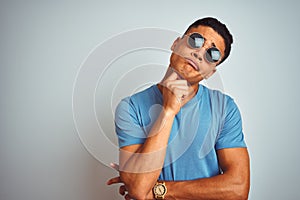Young brazilian man wearing blue t-shirt and sunglasses over isolated white background with hand on chin thinking about question,