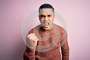 Young brazilian man wearing casual sweater standing over isolated pink background angry and mad raising fist frustrated and