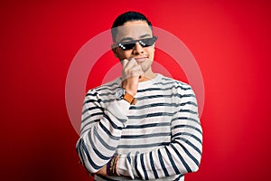 Young brazilian man wearing funny thug life sunglasses over isolated red background with hand on chin thinking about question,