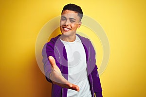 Young brazilian man wearing purple sweatshirt standing over isolated yellow background smiling friendly offering handshake as photo