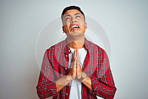 Young brazilian man wearing red shirt standing over isolated white background begging and praying with hands together with hope